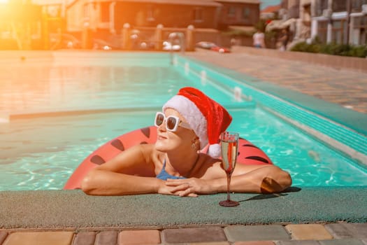 Woman pool Santa hat. A happy woman in a blue bikini, a red and white Santa hat and sunglasses poses near the pool with a glass of champagne standing nearby. Christmas holidays concept