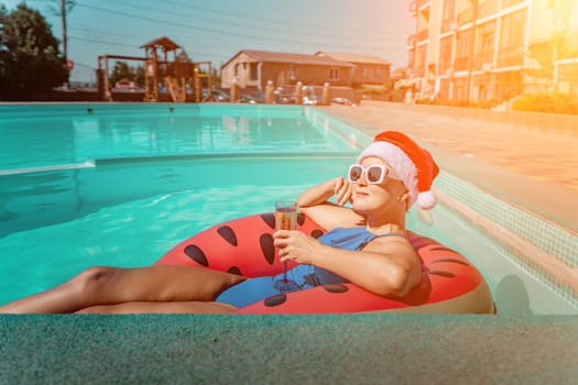 A happy woman in a blue bikini, a red and white Santa hat and sunglasses poses in the pool in an inflatable circle with a watermelon pattern, holding a glass of champagne in her hands. Christmas holidays concept