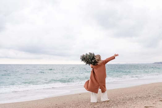 Blond woman Christmas sea. Christmas portrait of a happy woman walking along the beach and holding a Christmas tree on her shoulder. She is wearing a brown coat and a white suit