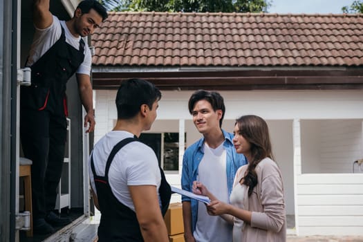 A satisfied couple signs the delivery checklist with assistance from professional movers after furniture handling. Employees in uniform ensure customer satisfaction. Moving Day Concept