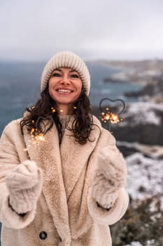 Outdoor winter portrait of happy smiling woman, light faux fur coat holding heart sparkler, posing against sea and snow background.