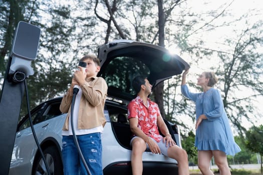 Little boy holding EV charger and point at camera with his family sitting on the trunk in background. Road trip travel with alternative energy charging station for eco-friendly car concept. Perpetual