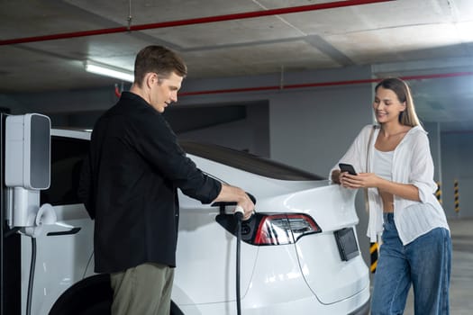 Young couple travel with EV electric car to shopping center parking lot charging in downtown city showing urban sustainability lifestyle by green clean rechargeable energy of electric vehicle innards