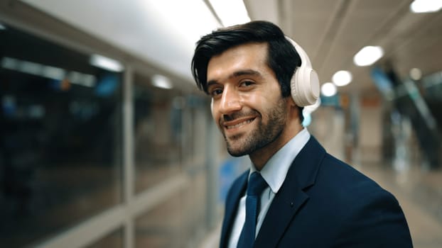 Closeup of smart business man wearing headphone and listening music while standing at train station. Professional executive manager looking at camera while waiting for train or subway. Exultant.