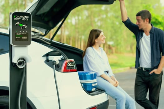 Lovely young couple recharging battery for electric car while relaxing and sitting on the trunk during road trip travel EV car in autumnal forest. Eco friendly travel on vacation during autumn. Exalt