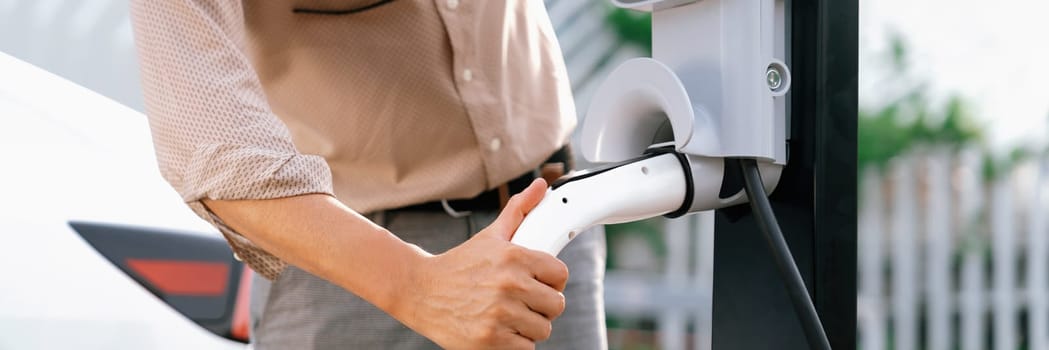 Young man recharge electric car's battery from charging station in city commercial parking lot. Rechargeable EV car for sustainable environmental friendly urban travel. Panorama Expedient