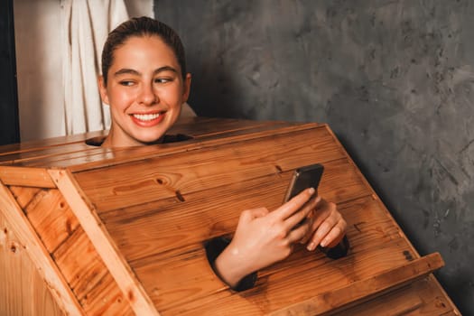 A portrait of gorgeous caucasian woman playing her mobile phone while using wooden sauna cabinet in warm tone. Attractive female with beautiful skin taking a photo. Gray background. Tranquility