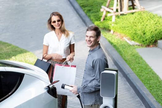 Young couple travel with EV electric car charging in green sustainable city outdoor garden in summer shows urban sustainability lifestyle by green clean rechargeable energy of electric vehicle innards