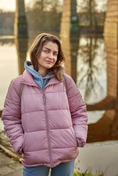 A young woman standing at the shore looking at the river in autumn sunny day. Street view, copy space for text, travel photo. Happy tourist woman on the bank of the river in autumn in warm clothes. Tourists enjoy their vacation, winter season. Romantic look and travel concept. A joyful mood in a Caucasian girl. Winter Wonderland: Enchanting Girl by the Riverside in Autumn.