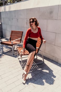 Portrait of a woman on the street. An attractive woman in glasses, a red blouse and a black skirt is sitting on a bench outside