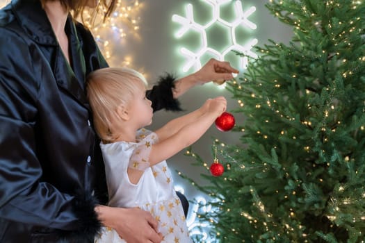 A mother with a 2-year-old daughter decorates the Christmas tree. Mom in a black suit, a girl in a white dress, her daughter hangs a red ball on the Christmas tree. Merry Christmas and New Year concept.