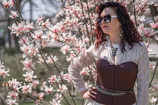 Magnolia park woman. Stylish woman in a hat stands near the magnolia bush in the park. Dressed in white corset pants and posing for the camera