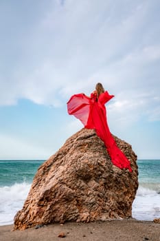 woman sea red dress. Woman with long hair on a sunny seashore in a red flowing dress, back view, silk fabric waving in the wind. Against the backdrop of the blue sky and mountains on the seashore