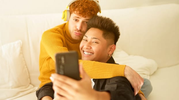 Gay couple embracing and smiling while taking selfie together sitting on the sofa