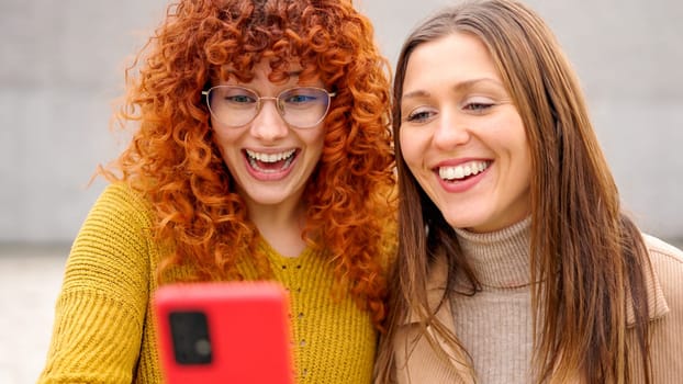 Two female friends laughing while using phone together in the street