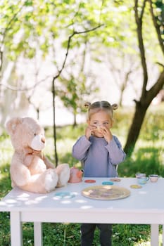 Little girl drinks tea from a cup with a teddy bear at the table at a tea party. High quality photo