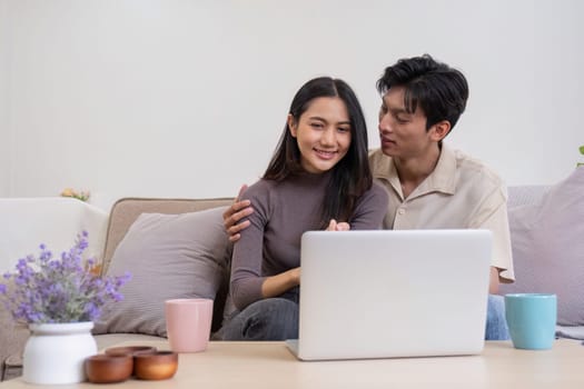 Asian couple watching movies on the internet using laptop at home Smiling Thai man and woman sitting on sofa, hugging each other, looking at computer screen. Surf the web together copy space.