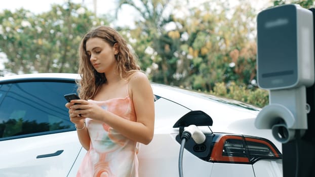 Young woman use smartphone to pay for electricity at public EV car charging station in nature. Modern environmental and sustainable automobile transportation lifestyle with EV vehicle. Synchronos