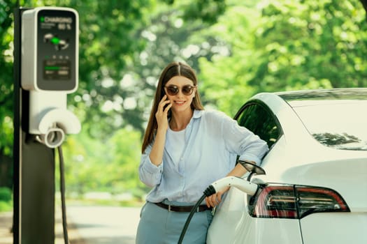 Young woman talking on smartphone while recharging electric car battery charging from EV charging station during vacation holiday road trip at national park or summer forest. Exalt