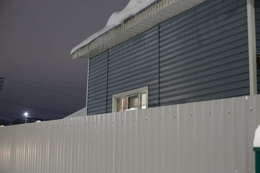 Photo in the evening, a private house behind a corrugated board fence. The color is gray. Twilight.