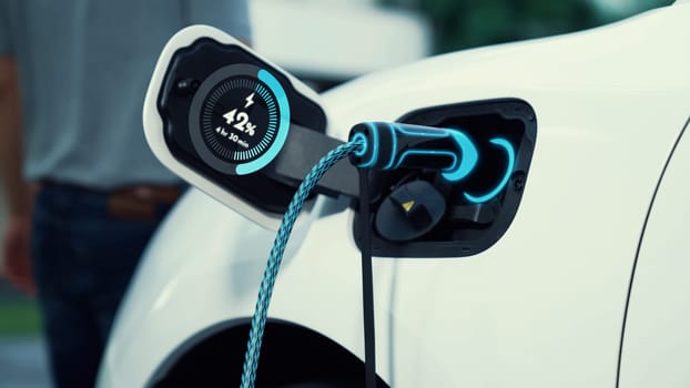 EV charger from home charging station plug in and recharging EV car display battery status hologram with blurred background of modern man walking. Smart futuristic home energy infrastructure. Peruse