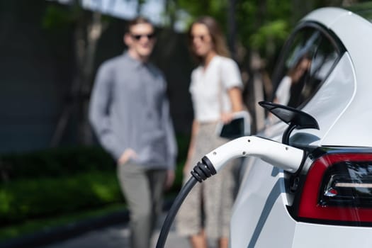 Young couple travel with EV electric car charging in green sustainable city outdoor garden in summer shows urban sustainability lifestyle by green clean rechargeable energy of electric vehicle innards