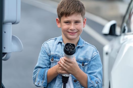 Road trip vacation with eco-friendly EV car and little boy pointing EV charger at camera. Electric vehicle and charging station using clean and sustainable energy for environment protection. Perpetual