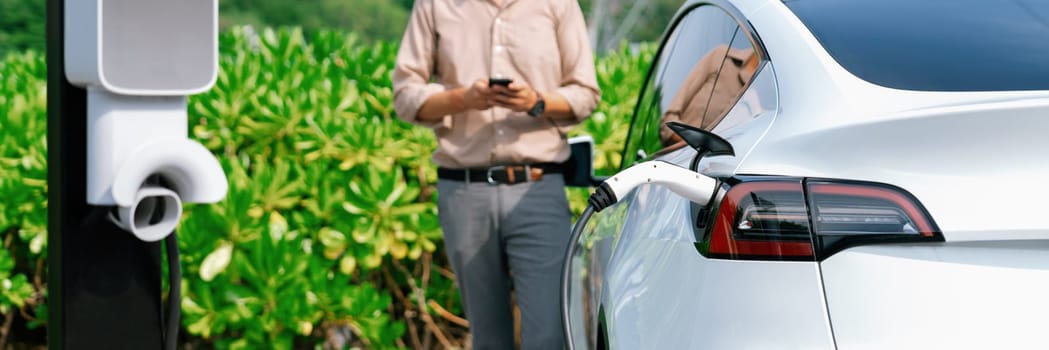 Young man use smartphone to pay for electricity at public EV car charging station green city park. Modern environmental and sustainable urban lifestyle with EV vehicle. Panorama Expedient