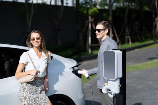 Young couple travel with EV electric car charging in green sustainable city outdoor garden in summer shows urban sustainability lifestyle by green clean rechargeable energy of electric vehicle innards