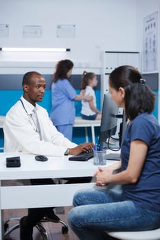 In medical office, African American doctor discusses with caucasian lady about healthcare and medicine. Black man using the desktop PC, talks to the patient while wearing a lab coat and mask.