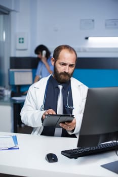 Using a tablet, male doctor reads patient records. Caucasian man in a lab coat is reviewing medical data on a desktop computer and digital device in the clinic office.