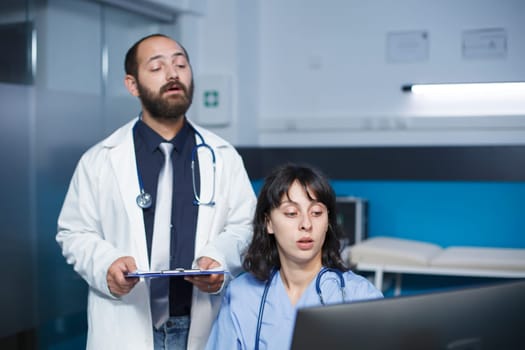 Medical professionals discuss patient care and uses a tablet and computer. Male physician in lab coat gestures as he talks with female nurse. Digital technology and medical data support collaboration.