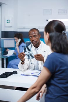 African American female specialist reviewing x-ray image and analyzing imaging results. Radiology expert examines radiography scan and provides diagnostic results to caucasian woman patient.