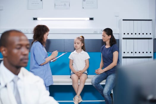 Image shows a mother and daughter seated on a hospital bed, receiving medical consultation from a female nurse practitioner. Communication with patient regarding health. Background focus.