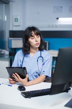 For healthcare, a medical assistant uses a computer and tablet with a touch screen. Working late at night, a female nurse holds a gadget and reviews patient consultations. Expert in electronic devices