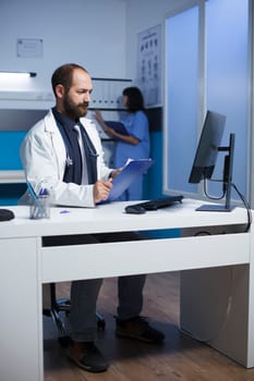 At the clinic, committed male physician is checking and reviewing his healthcare notes. Using a desktop monitor and clipboard, a doctor prepares for patient medical consultations.