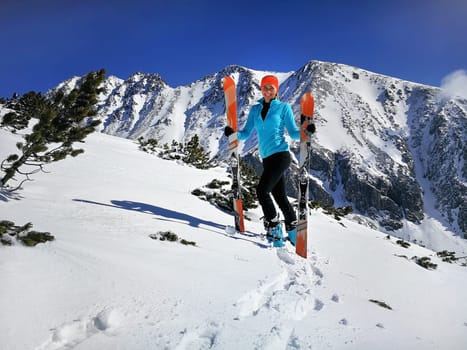 Young woman in light blue jacket standing, smiling happy holding with mountaineering ski and poles in hands, sun shines on snow, bright sky over mountains behind