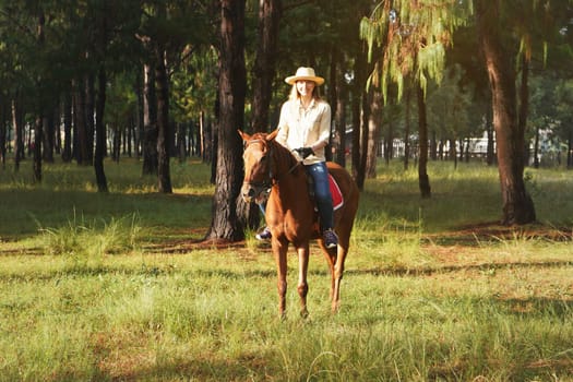 Young woman in shirt and straw hat, rides brown horse in the park, blurred background with trees