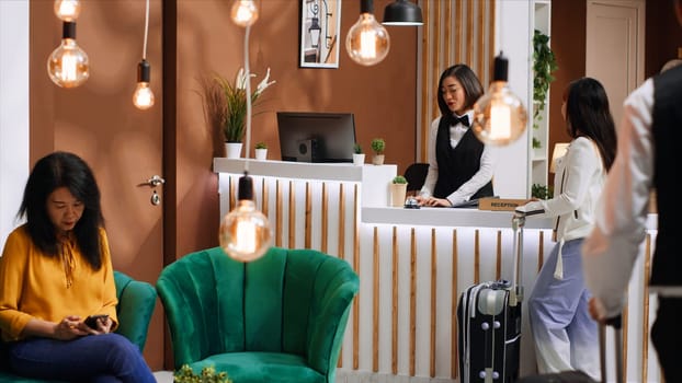 Young traveler arriving at hotel lobby with luggage, receptionist asking guest about her room reservation for registration. Employee welcoming client at five star resort, hospitality industry.