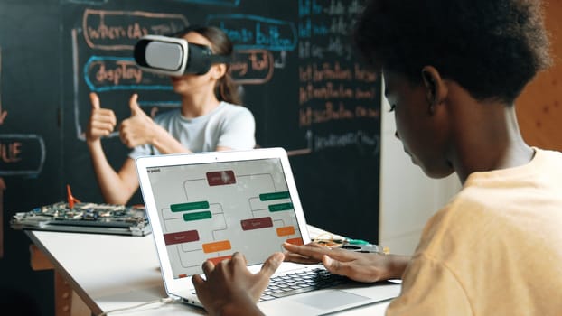Caucasian school girl wear VR glass and showing thumb up while african boy working on laptop at blackboard coding engineering prompt written in STEM class. Creative education concept. Edification.
