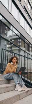 Stylish female entrepreneur working on laptop sitting outside on modern building background