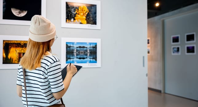 Young person at photo frame write digital book leaning against at show exhibit artwork gallery, Asian woman holding tablet at art gallery collection in front framed paintings looking pictures on wall