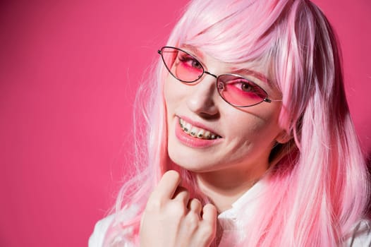 Close-up portrait of a young woman with braces in a pink wig and sunglasses on a pink background