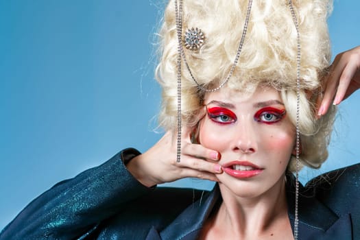 Portrait of funny princess with vintage wig and red makeup making faces. Woman wearing elegant jacket on blue background. Royal party in blue studio.