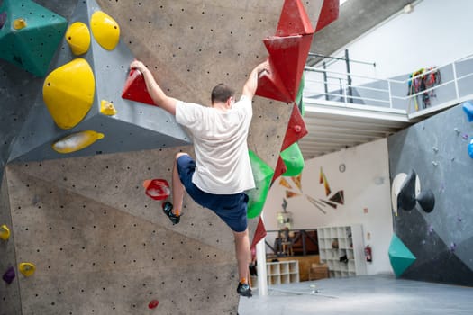 Rear view of adult man climbing wall at sports center. Full length of active male athlete practicing bouldering in gym. Sportsman climbing artificial rock without safety rope.