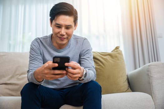 Asian older middle aged man relaxing sitting on sofa using smart mobile phone at home, Happy mature men smiling chatting with social media on smartphone in living room, holding device