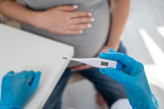 Pregnant woman with fever at doctor's appointment. Therapist holds an electronic thermometer with a temperature of 37.5