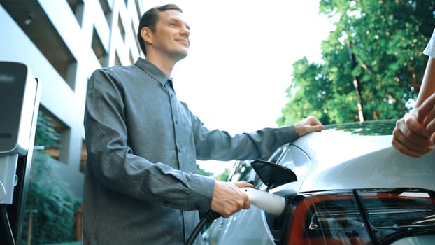 Young couple travel with EV electric car charging in green sustainable city outdoor garden in summer shows urban sustainability lifestyle by green clean rechargeable energy of electric vehicle innards