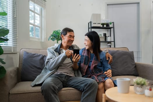 Retired elderly couple sits on couch using mobile together and relax in their home. Senior Activity Concept.