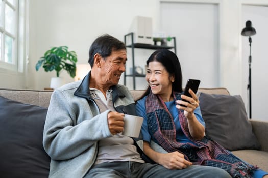Retired elderly couple sits on couch drink tea and using mobile together and relax in their home. Senior Activity Concept.